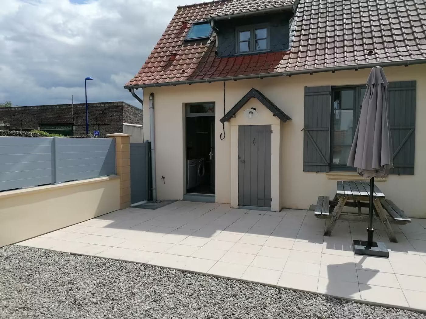 Façade de La Quiétude, maison de vacances au Crotoy : toit en tuiles, lucarne, volets gris et terrasse dallée devant la cour close